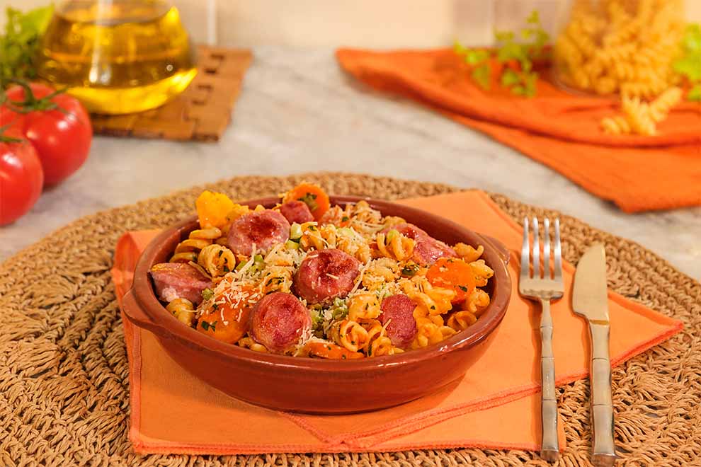 macarrão de panela de pressão com linguiça dentro de um pequeno bowl de porcelana marrom em cima de um guardanapo laranja e em cima de um jogo americano redondo de palha com talheres ao lado