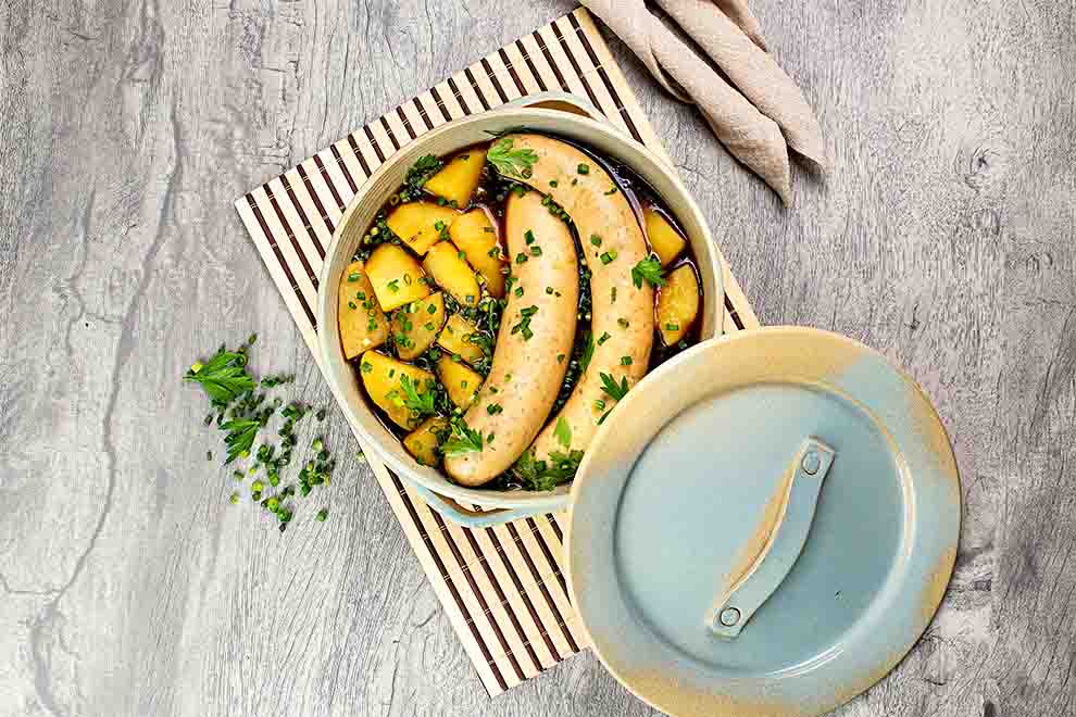 ensopado de salsicha frankfurt com batata e cheiro verde dentro de uma panela azul claro em cima de um pequeno jogo americano de bambu e em cima de uma mesa