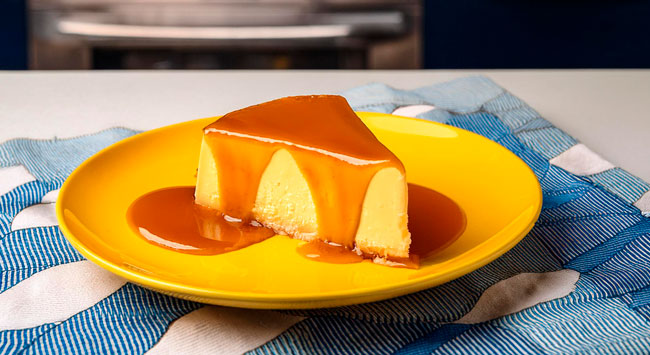 Fatia de pudim de leite condensado com calda caramelizada em prato amarelo sobre toalha listrada.