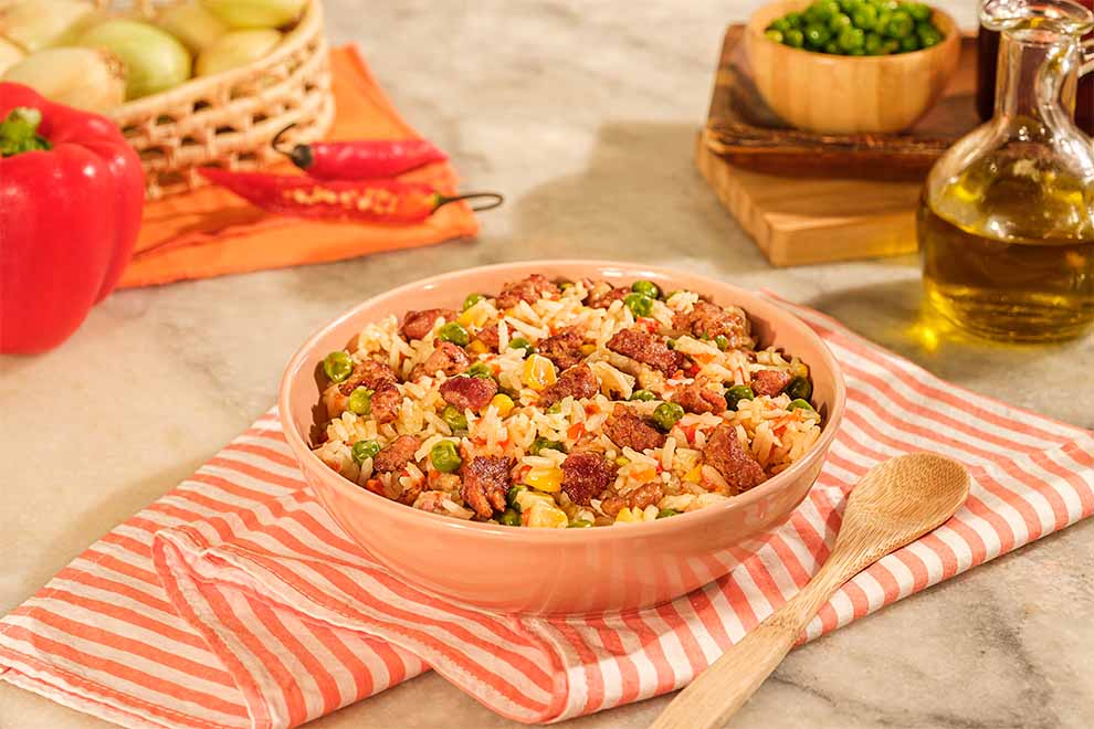 arroz com linguiça toscana dentro de um prato fundo de porcelana rosado com uma colher pequena de pau ao lado, em cima de uma mesa com um pano listrado em branco e alaranjado