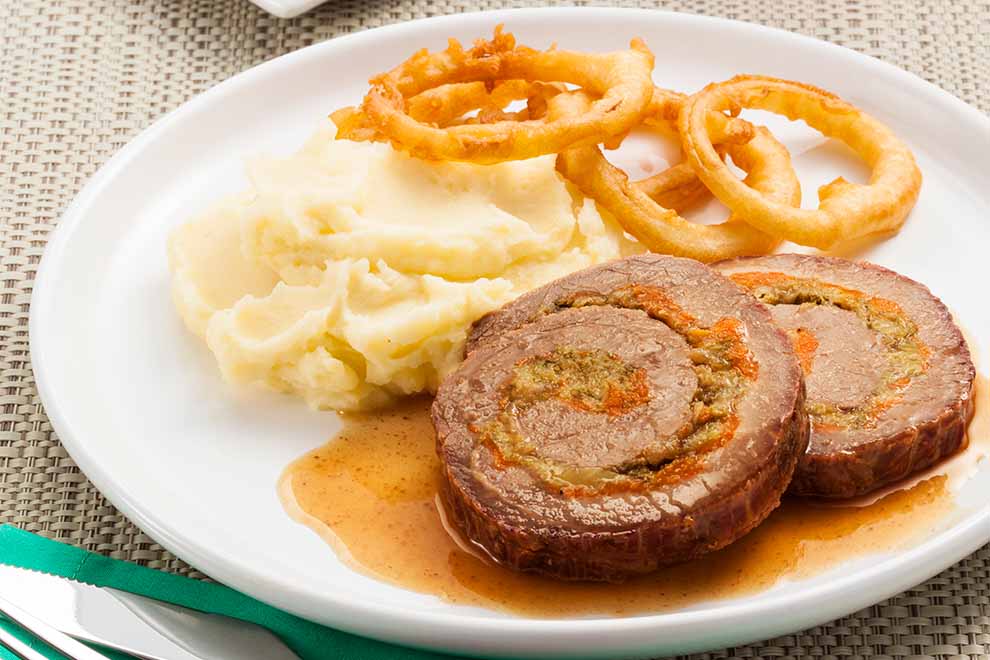 lagarto recheado com molho, purê e anéis de cebola dentro de um prato branco de porcelana em cima de um jogo americano bege com talheres ao lado