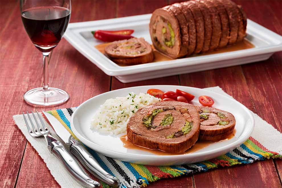 lagarto recheado com calabresa e legumes dentro de um prato branco de porcelana e em cima de um pano listrado com talheres ao lado em cima de uma mesa de madeira escura