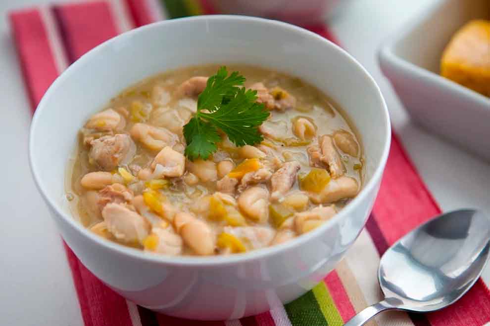 chilli de frango com feijão branco dentro de um pequeno bowl branco de porcelana e uma colher ao lado em cima de um pano listrado
