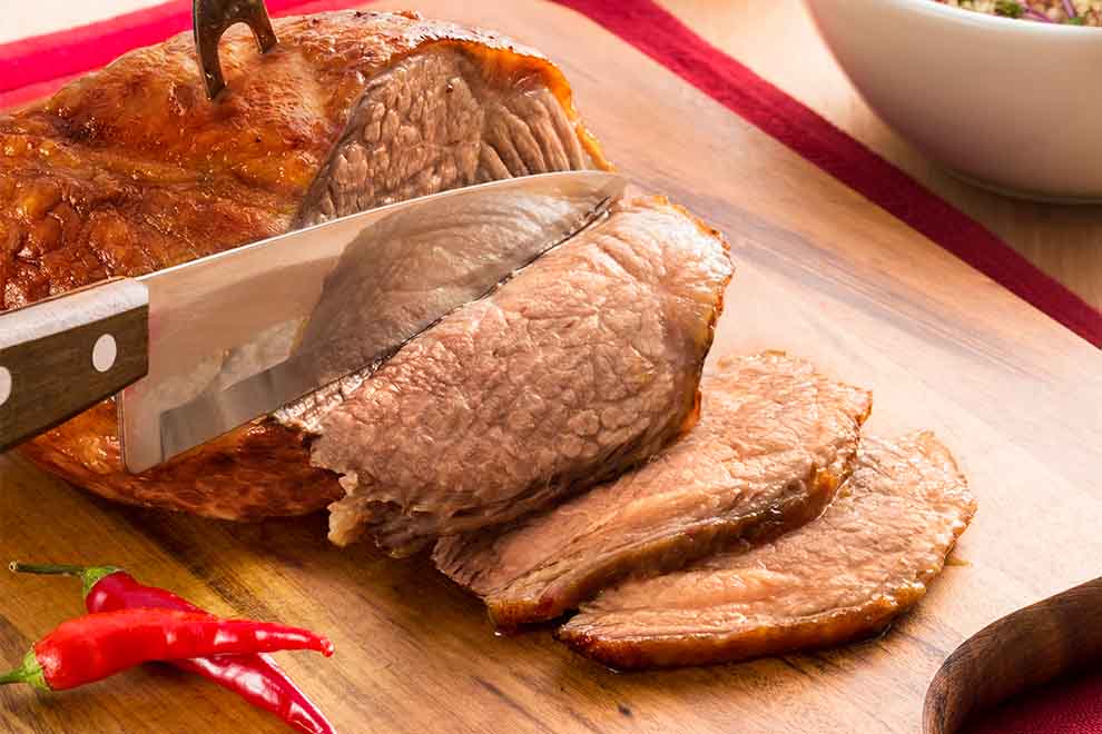 cupim assado na churrasqueira sendo cortadas em fatias em cima de uma tábua de madeira com pimentas ao lado e em cima de uma pano vermelho em cima de uma mesa de madeira
