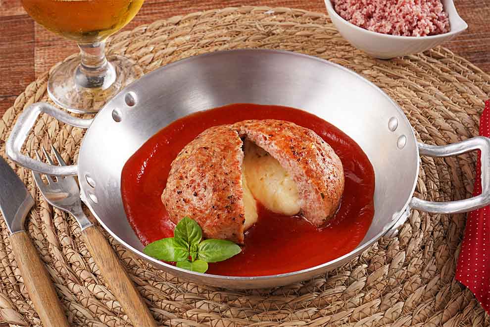 polpetone de mortadela dentro de uma pequena panela de alumínio, com talheres ao lado e em cima de um jogo americano em palha