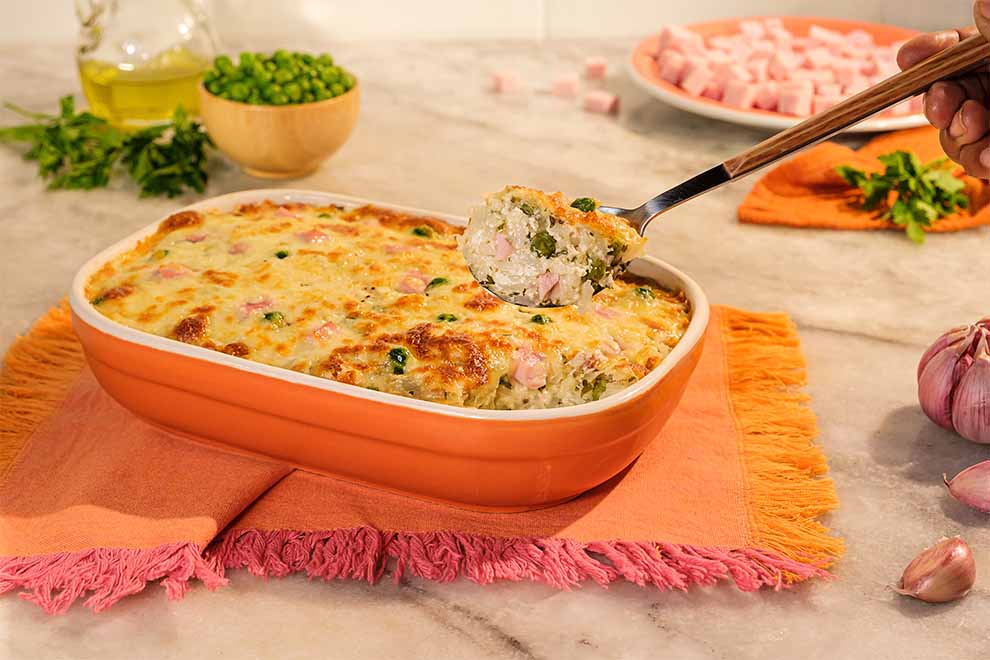 arroz de forno levissímo e queijo dentro de uma travessa de porcelana alaranjada em cima de uma mesa e um pano alaranjado e uma mão segurando uma colher