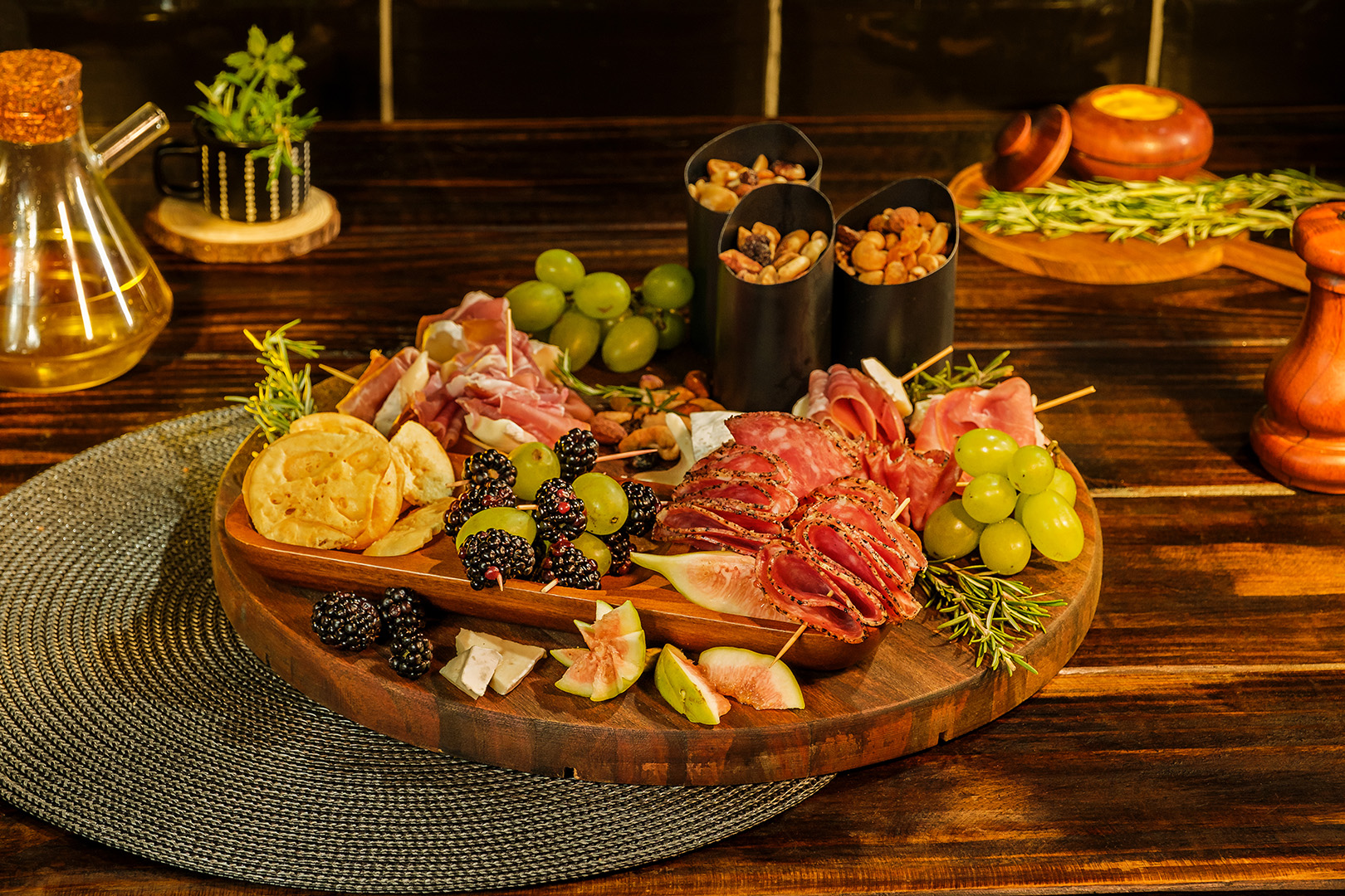 Tábua de frios com salames, figos, uvas, castanhas em cumbucas, amora, e presunto cru arrumados em tábua de madeira.