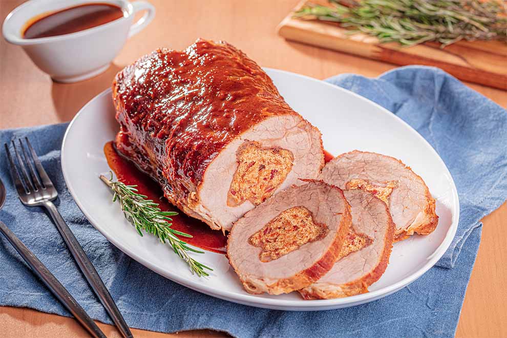 lagarto recheado com farofa defumada dentro de um prato branco de porcelana em cima de um pano azul com talheres ao lado em cima de uma mesa de madeira