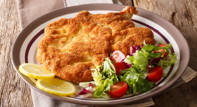 Bife à milanesa servido com salada de aflace, cebola roxa e tomatinhos em um prato raso redondo cumum, com rodelas de limão siciliano.