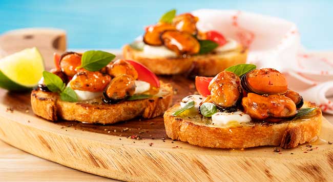 Bruschettas de pão italiano com mexilhões cozidos, mozzarella de búfula, tomate e manjericão servidas em uma tábua de madeira.