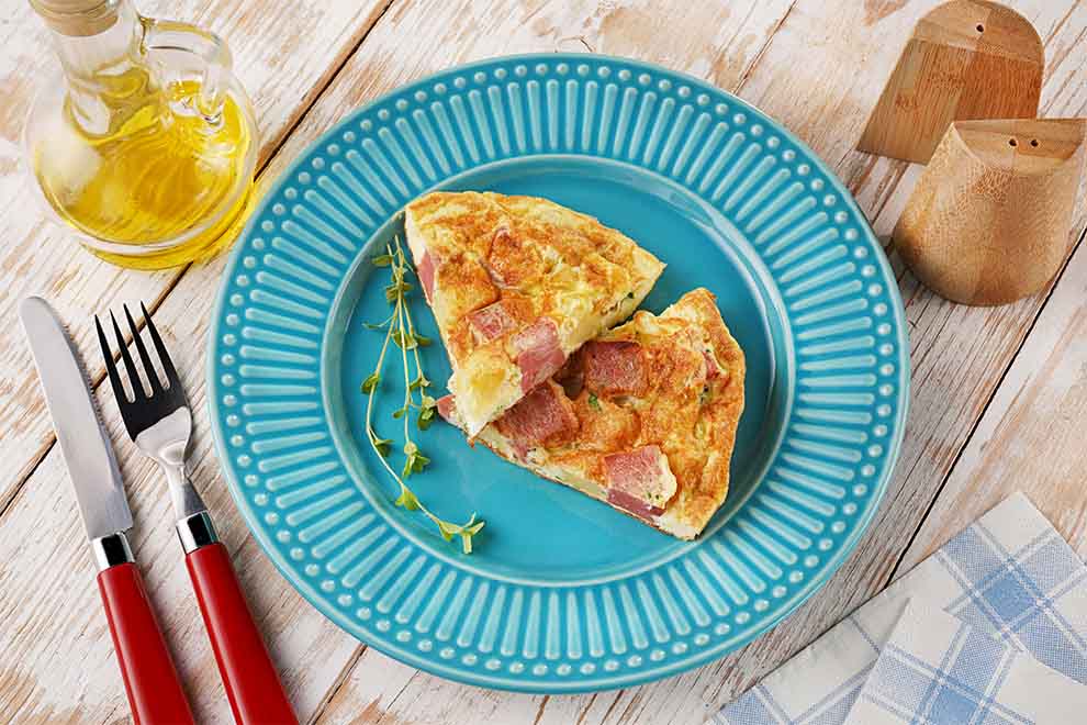fritada espanhola de mortadela dentro de um prato azul de porcelana com talheres ao lado e um guardanapo xadrez em azul e branco em cima de mesa de madeira