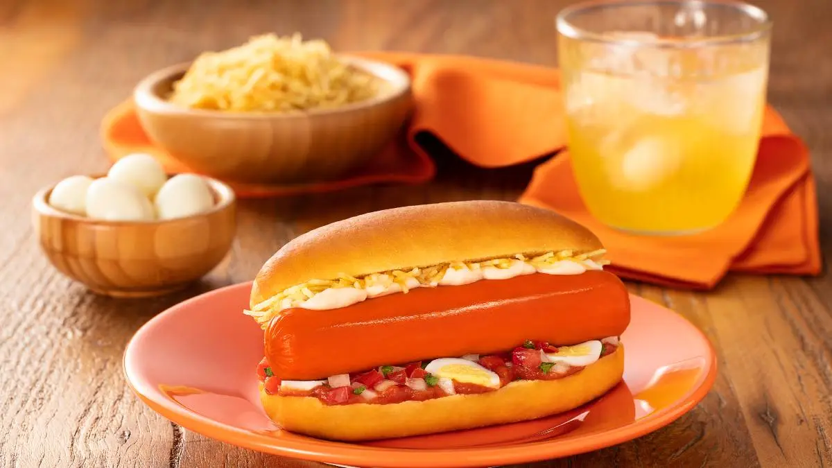 A imagem mostra um cachorro-quente servido em um prato laranja. O pão está levemente tostado e recheado com uma salsicha, maionese, batata palha, ovos de codorna cortados e vinagrete (uma mistura de tomate, cebola e ervas). Ao fundo, desfocados, estão uma tigela com batata palha, uma tigela com ovos de codorna inteiros e um copo de bebida amarela com gelo, sobre uma mesa de madeira. Há também um guardanapo laranja ao fundo, complementando a cena.
