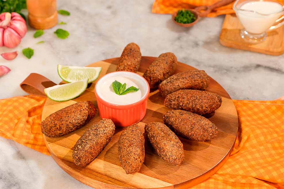 kibe frito em cima de uma tábua redonda de madeira com um pequeno bowl com molho dentro e dois pedaços de limão ao lado, em cima de uma mesa e um pano alaranjado