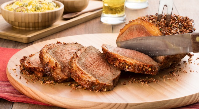 Churrasco de picanha sendo fatiada em uma tábua de madeira redonda clara. Atrás da tábua, alguns potes de molhos para acompanhar.