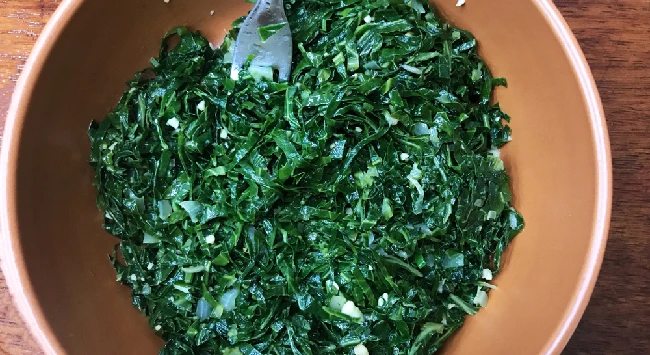 A imagem mostra uma frigideira com couve fatiada e refogada com azeite e alho finamente picado. A couve parece estar macia e tem uma cor verde-escuro. A frigideira é rosada e está posicionada sobre uma tábua de madeira marrom. Há um garfo de metal puxando um pouco de couve fatiada.