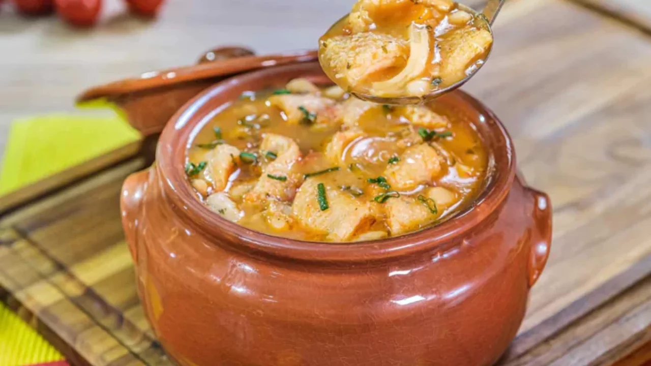 A imagem apresenta uma porção de dobradinha servida em uma panela de barro, típica da culinária brasileira. Na imagem, o caldo tem uma cor dourada, indicando o uso de especiarias e temperos. Um detalhe adicional é a colher que levanta um pedaço da dobradinha, mostrando a textura do prato.