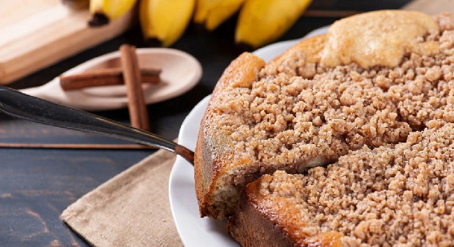 Cuca de banana servido em prato branco com bananas inteiras atrás.