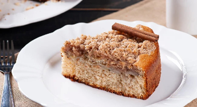 Fatia de cuca de banana servida em prato branco com canela em pau em cima.