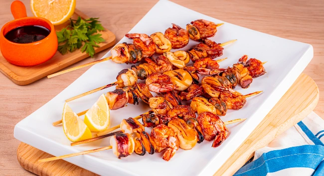 Espetos de camarão e lula grelhados com limão e molho escuro, servidos com toque rústico e colorido. Uma refeição fresca e irresistível!