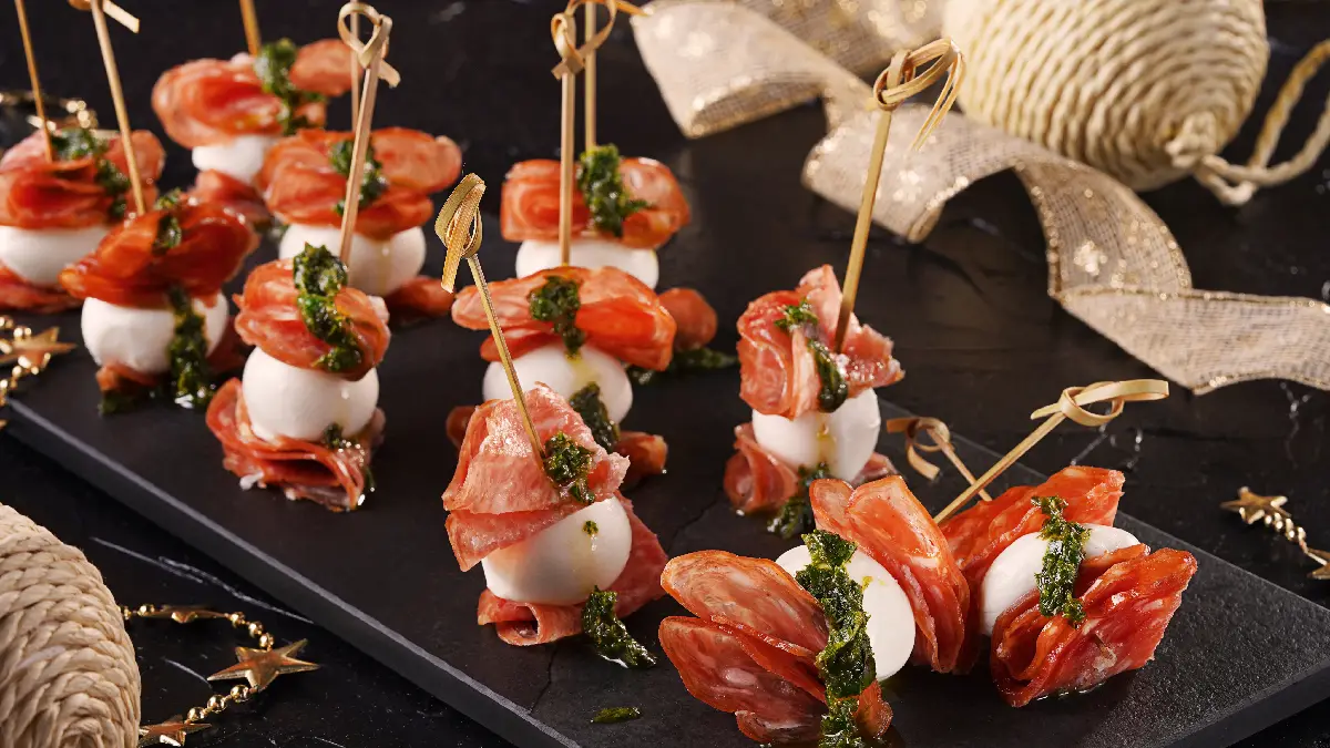 Espetinhos de salame com muçarela de búfula e pesto de manjericão em uma tábua preta.
