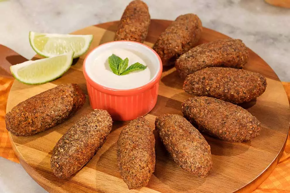 Diversos kibes fritos em cima de uma tábua redonda de madeira, com uma fatia de limão e um combuca de molho, sob uma mesa de mármore