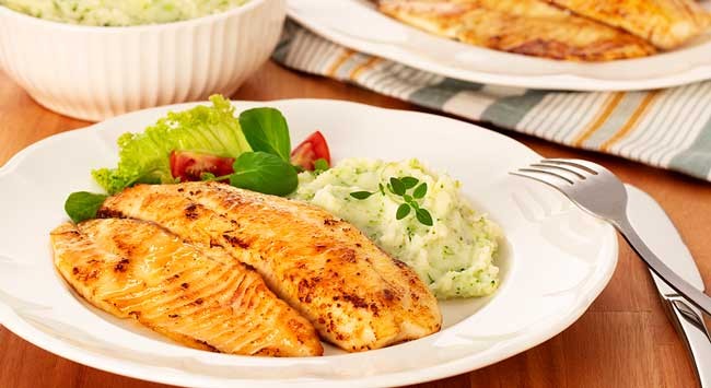 Mesa posta com Filé de Tilápia Grelhada e Purê de Batatas e Brócolis servidos em um prato redondo branco. Atrás, um outro prato com filés de tilápia grelhados e, do lado esquerdo, o restante do purê de batatas e brócolis servido em um ramequim redondo e branco.