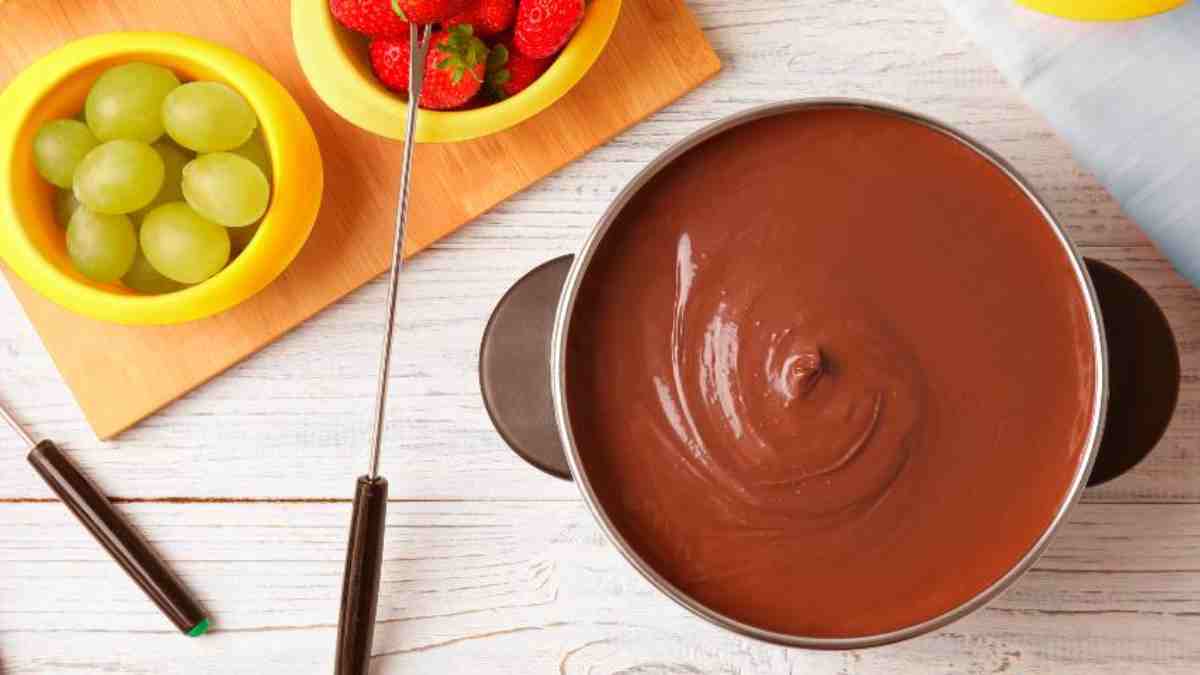 Panela de fondue de chocolate acompanhado de tábua amarela com dois potinhos servindo frutas (morangos e uvas) separadamente e dois garfos de fondue ao lado.