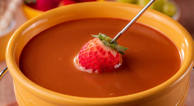 Uma tigela de fondue de doce de leite. A tigela é de cerâmica, de cor amarela, e está cheia de doce de leite cremoso. Um morango vermelho e fresco está parcialmente mergulhado no fondue, espetado por um garfo longo de metal. Ao fundo, fora de foco, há frutas adicionais, incluindo uvas e morangos, sugerindo que estão prontas para serem mergulhadas no fondue.