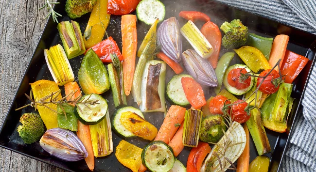 Travessa com legumes assados como abóbora, pimentão vermelho, cebola roxa e cenoura, finalizados com ervas frescas picadas.