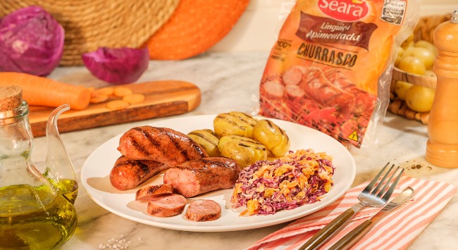 A imagem destaca um prato branco redondo com linguiça de churrasco fatiada, batatas assadas e salada coleslaw. O prato está sobre um pano listrado e atrás, do lado direto, há uma embalagem de linguiça apimentada churrasco Seara.
