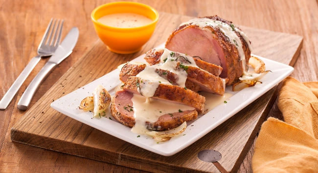 Lombo suíno ao creme de requeijão servido em uma travessa branca. Ao fundo, uma tigela amarela com creme.