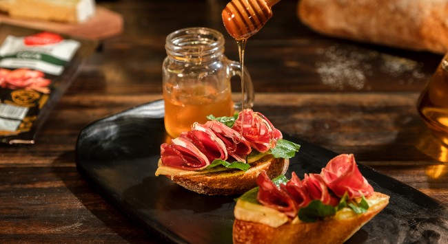 Duas unidades de Bruschetta de copa com queijo brie em um prato retangular preto. Há um pote de mel ao lado e um utensílio despejando mel por cima do aperitivo. No fundo, do lado direito, um embalagem de copa fatiada seara gourmet e uma faia de queijo brie. Do lado direito, um pão ciabatta inteiro.