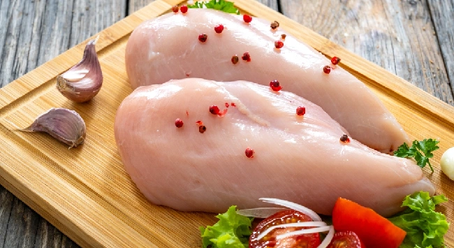 Dois pedaços de peito de frango cru em uma tábua de madeira com grãos de pimenta, alho e folhas de ervas.