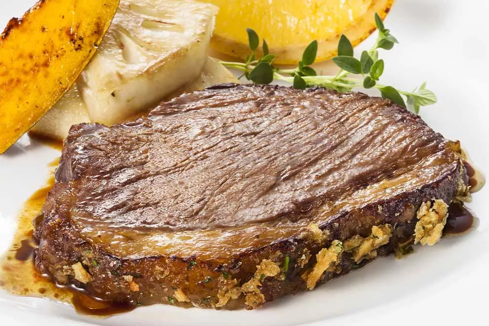 Imagem de uma fatia de picanha feita na chapa, com uma batata frita, pedaços de abacaxi assado e uma fatia de limão siciliano ao lado, sob uma prato redondo branco, em cima de uma mesa branca