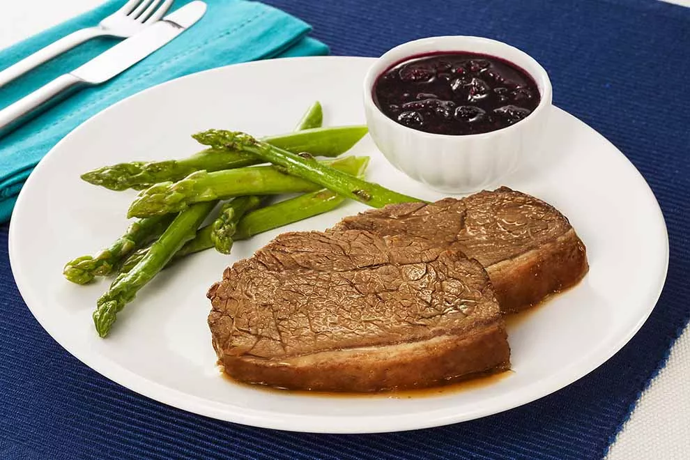 Imagem de duas fatias de picanhas feitas na chapa, com aspargos cozidos e uma combuca de frutas vermelhas ao lado, sob um prato redondo branco, com um par de talheres ao lado, em cima de pano azul, sob uma mesa branca