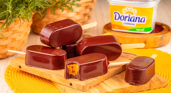 Picolés de bolo de cenoura cobertos com chocolate sobre uma tábua de madeira, ao lado de uma embalagem de margarina Doriana.