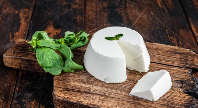 Ricota inteira com um pedaço cortado servida sobre tábua rústica de madeira, acompanhada de folhas frescas de manjericão ao lado.