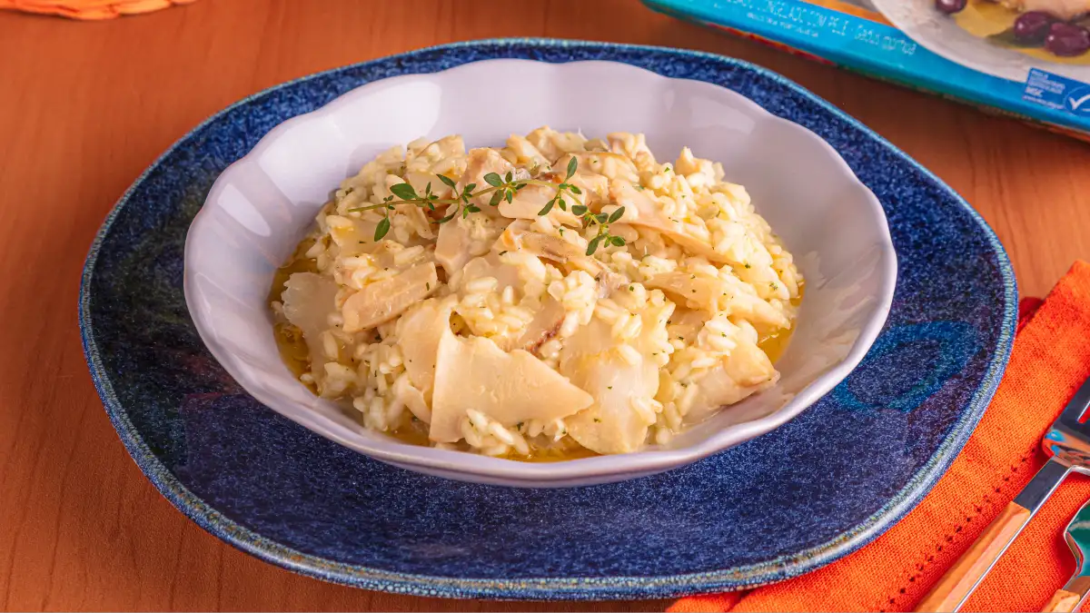 Prato de risoto de bacalhau com lascas de peixe e ervas frescas em um bowl branco, sobre um prato azul, com guardanapo laranja ao lado.