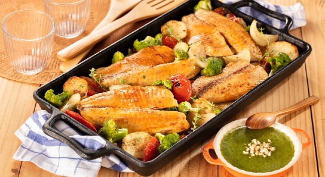 Travessa preta com filés de tilápia e uma salada diversa de legumes acompanhada de molho pesto.