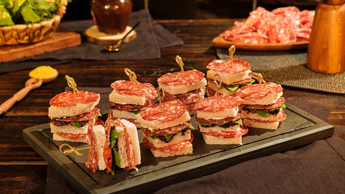 Imagem destaca 8 espetinhos de torta fria no palito feitos com pão de forma, salame fatiado, creme de ricota com curry, geleia de ameixa e espinafre: uma das receitas de petiscos frios para comer em dias quentes.